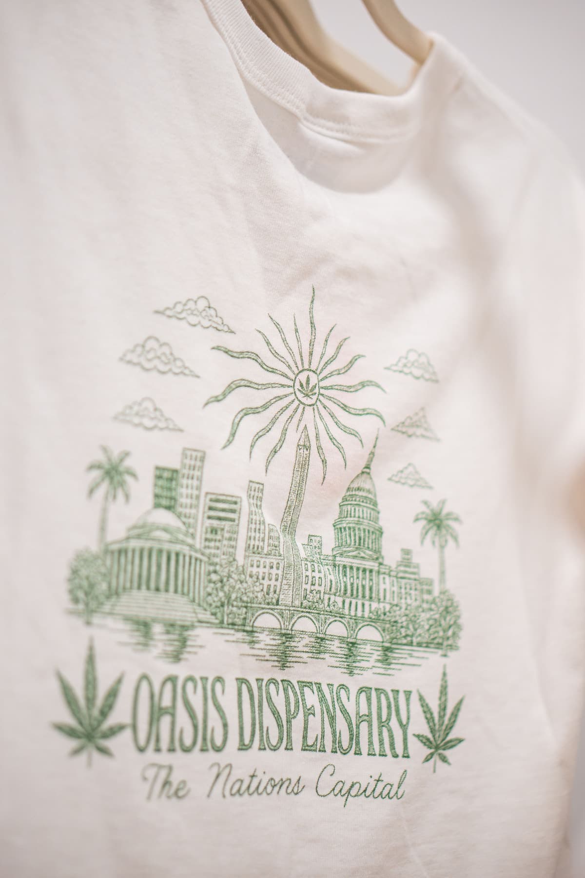 A cream Oasis Dispensary T-shirt printed with an illustrated Washington, DC skyline